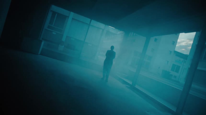 A lone figure stands in a hazy urban space under blue light