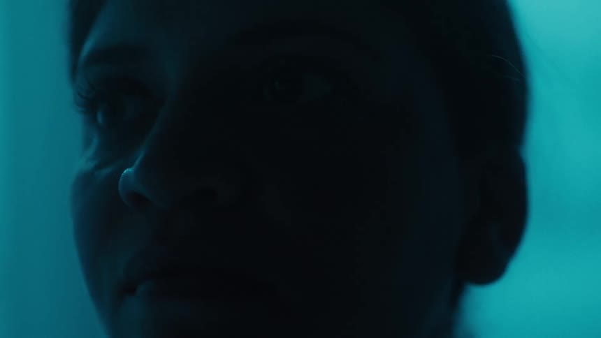 A dimly lit closeup shows the profile of a person with their head tilted slightly down the face is mostly in shadow against a blue background and their features are barely visible