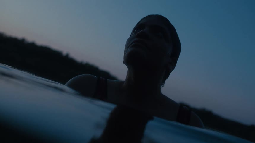 A woman bathing in a body of water as night falls only her head and shoulders out as she looks toward the sky