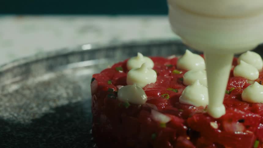 A hand squeezing a thick white sauce from a plastic bottle onto a finely chopped red vegan dish garnished with small white and green ingredients