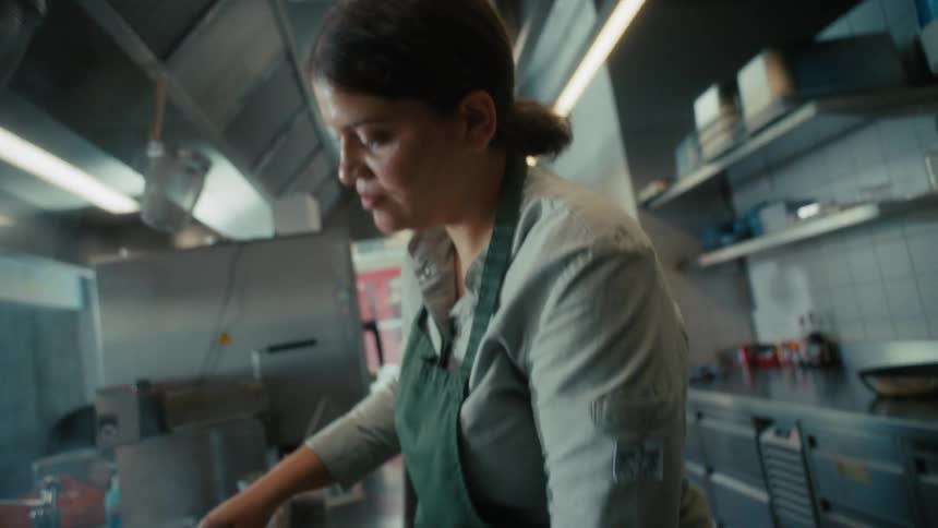 A side shot of a woman with her hair tied back focusing intently on her work in a professional kitchen setting