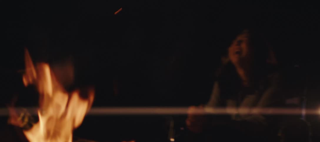 A group of friends sits by a campfire at night smiling and talking