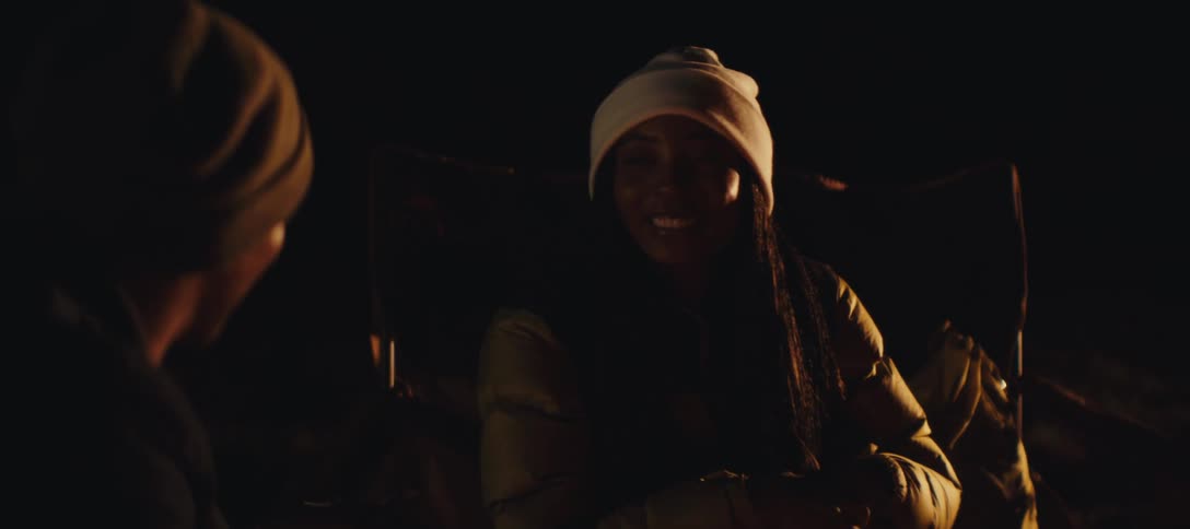 Woman wearing a beanie smiles while sitting by the campfire at night