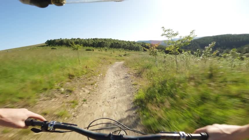 A cyclist rides down a bumpy dirt trail with the handlebars and hands of the rider visible in the foreground and a wide green and hilly landscape in the background