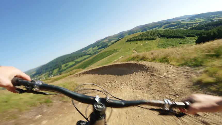 A firstperson pointofview shot shows someone riding a mountain bike downhill on a dirt trail with the handlebars and hands in the foreground and a vast scenic landscape of rolling green hills in the background