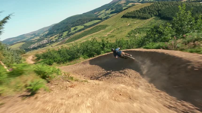 A mountain biker rides a winding dirt path down a large green hill with a village visible in the valley below