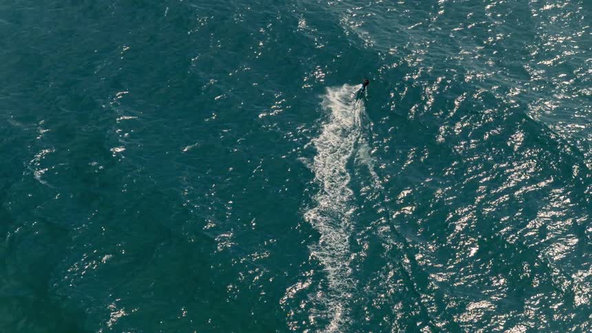 A wide overhead view shows a person in a black wetsuit riding a surfboard on a dark blue body of water leaving a trail of white foam behind them