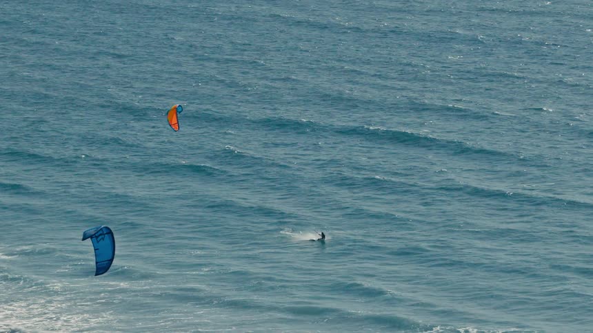 An aerial view captures two kiteboarders on the wavy blue ocean one person with an orange kite is riding the waves while a second kiteboarder with a blue kite is visible to the left