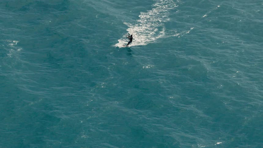 A surfer wearing a dark wetsuit rides a surfboard across a dark blue body of water leaving a trail of white foam behind him
