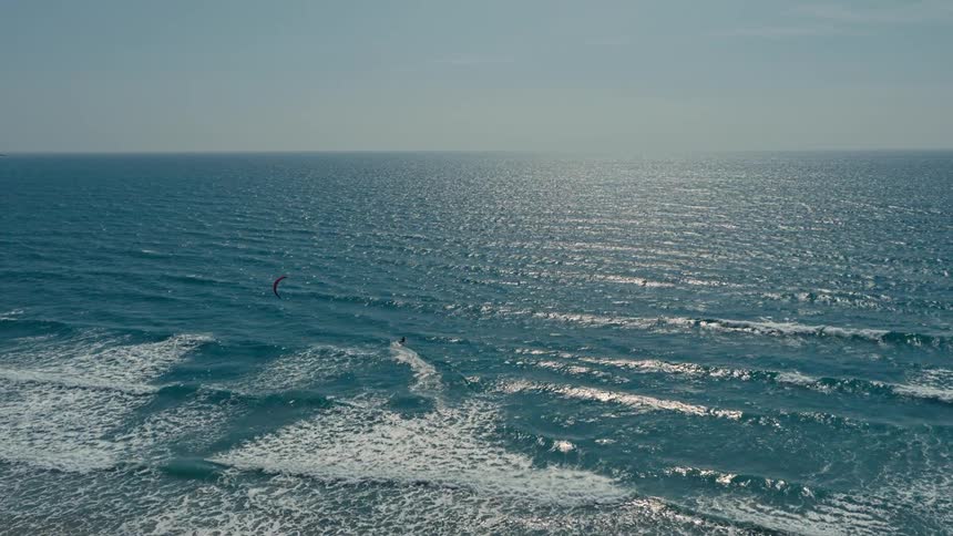 A lone kite surfer rides the waves on a bright clear day with the sun shimmering on the expansive blue ocean
