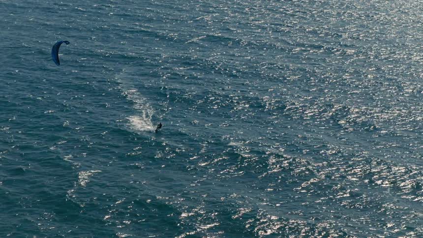 A lone kite surfer rides across a dark blue body of water leaving a thin white wake behind him as a blue kite flies above him