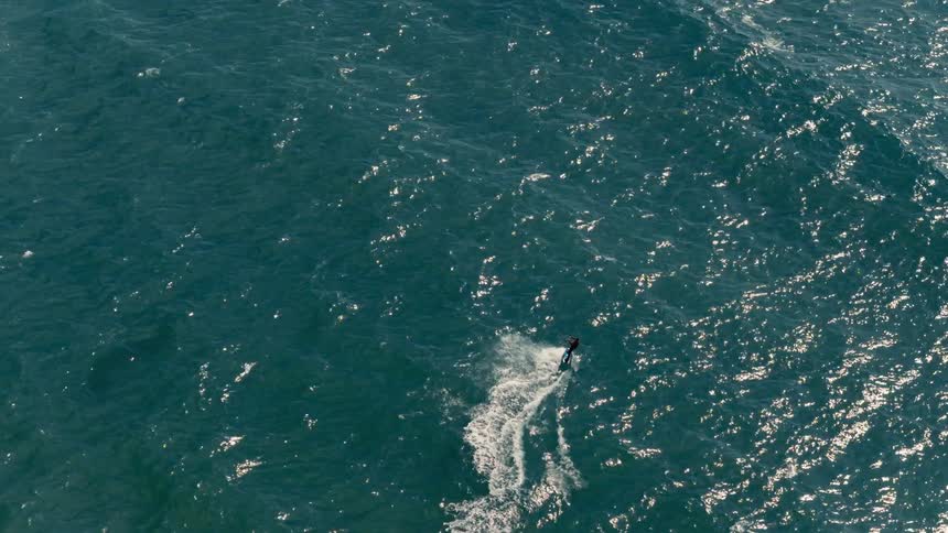 An aerial shot captures a kiteboarder carving through the deep blue ocean the person and their board are small but a distinct white wake follows them