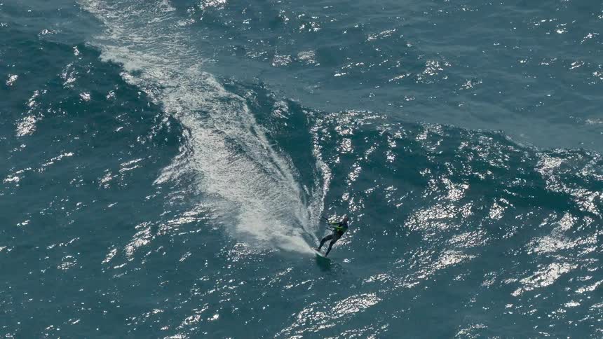 An aerial shot follows a person kiteboarding on the wavy blue ocean they are riding on a large wake creating a large foamy trail behind their board as they move