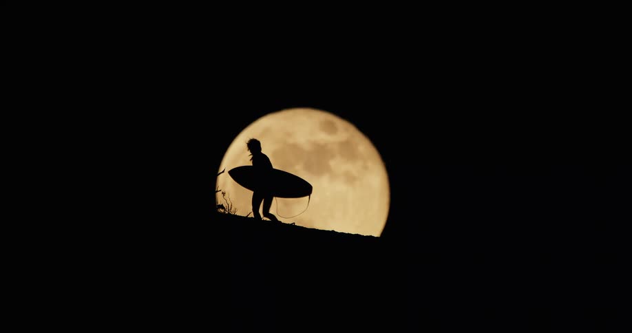 A surfer is seen in silhouette walking up a small hill while carrying a surfboard all set against the backdrop of a large full moon rising in the dark sky