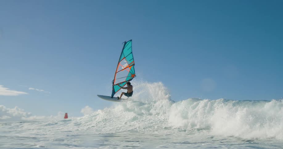 A person on a sailboard with a large orange and blue sail rides a foaming white wave on a body of water under a clear blue sky