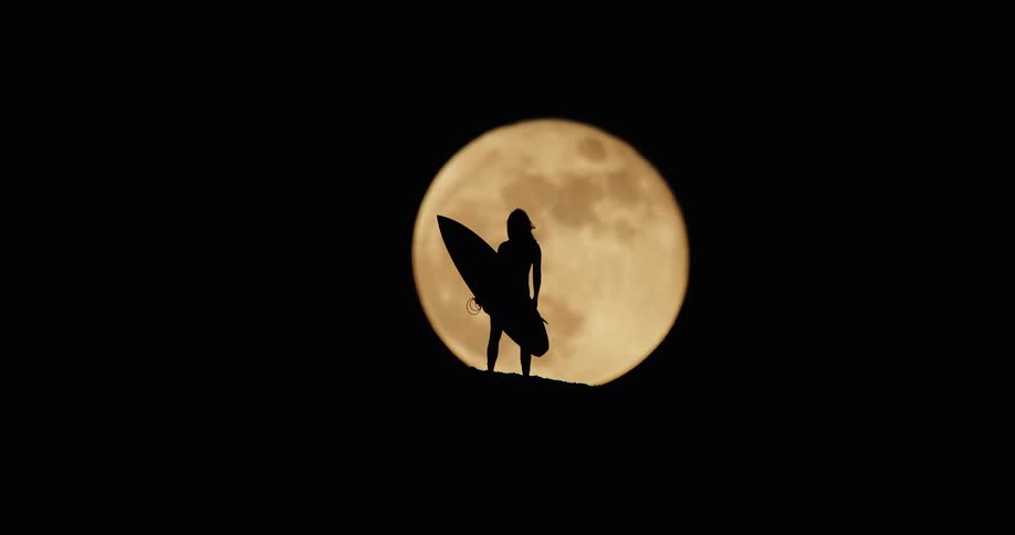 A silhouetted person is seen standing on a hill with a surfboard in hand with a large bright full moon directly behind them the person is completely dark against the luminous moon and sky