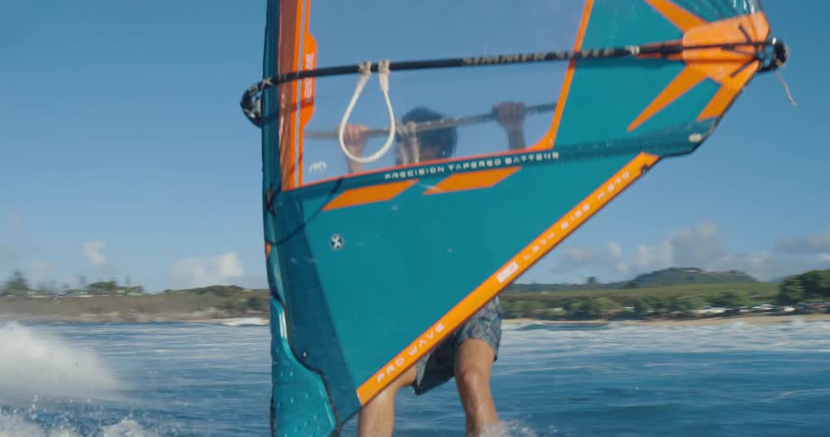 A man windsurfs on the ocean with a view of a grassy shore in the background the lowangle shot captures him expertly navigating the waves with his large blue and orange sail
