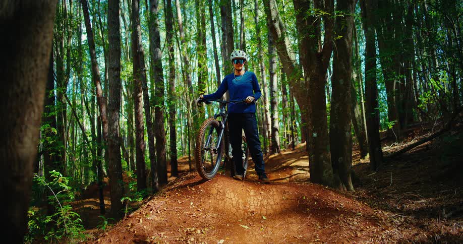 A wide shot captures a female mountain biker standing on a dirt mound in a dense forest she wears a helmet and glasses and stands next to her bike looking off to the right