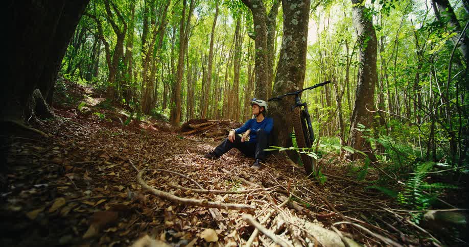 A woman in a bike helmet and blue shirt sits on the ground in a sundappled forest leaning against a tree her mountain bike is propped up next to her and she looks out into the trees as she rests