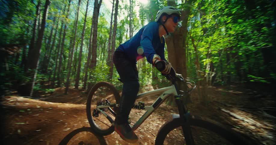 A dynamic shot captures a person on a mountain bike riding on a dirt track through a dense forest the rider comes down a small mound toward the camera then turns away to continue down the trail
