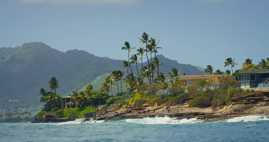 A scenic shot of a tropical coastline shows waves of deep blue water crashing against a rocky shore with palm trees and houses sitting on a lush hillside below a distant mountain range