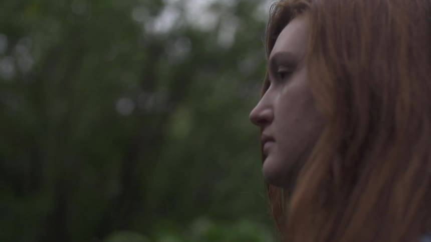 A tight shot of a young woman with red hair walking past trees
