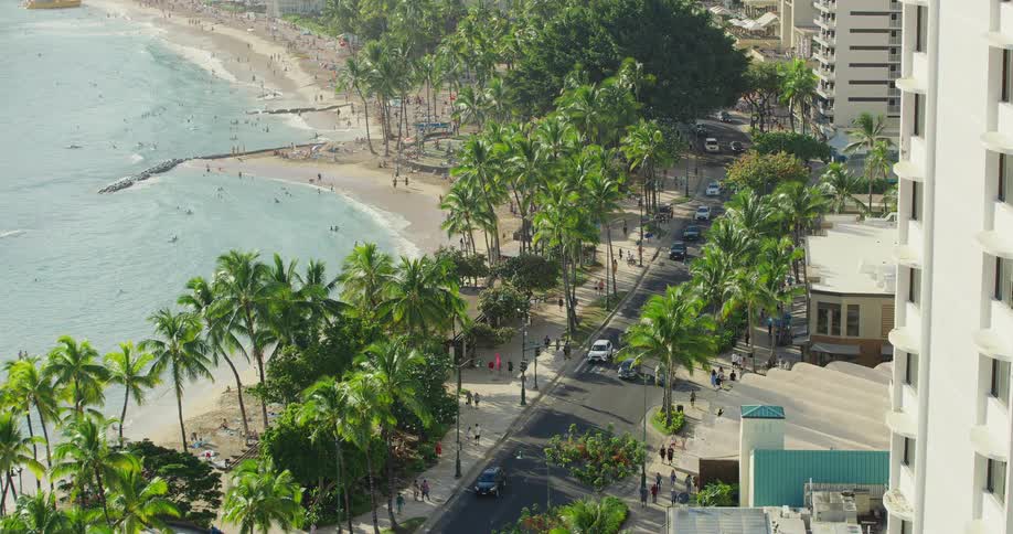 An elevated view of a vibrant tropical beach scene showing a busy street flanked by palm trees with a sandy beach and ocean on one side and tall buildings on the other