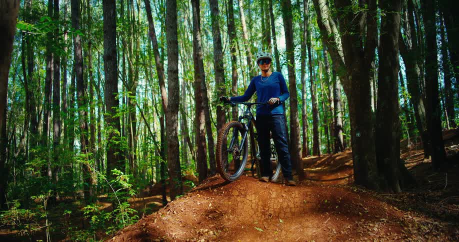 A person stands on a dirt mound in a dense forest holding a mountain bike they are wearing a helmet and a blue shirt and are looking into the distance