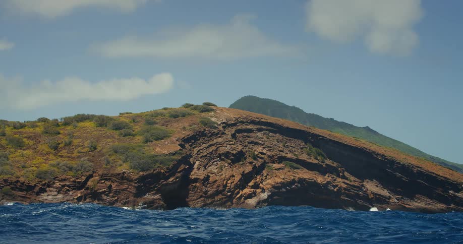 A shot from the waters surface shows a boat in the middle of a large body of water with waves rolling in front of it a large rocky cliff covered with green vegetation rises in the background