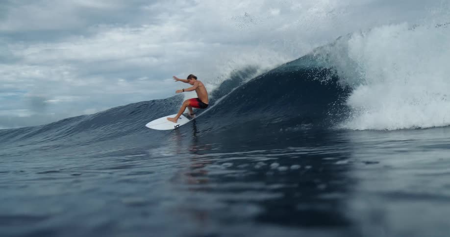 A shirtless man is surfing on a large ocean wave he is wearing red shorts and balancing on his surfboard as the wave curls behind him