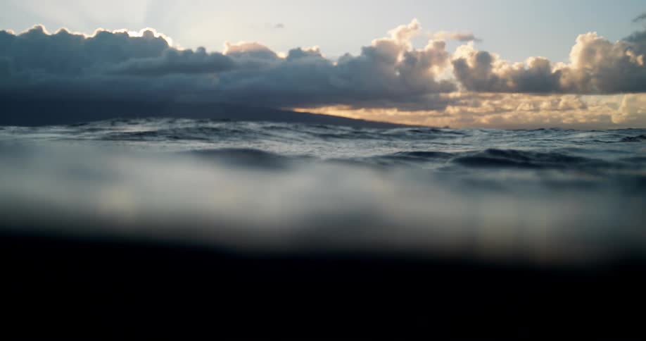 The shot captures the surface of the ocean at sunset with dark clouds and golden light breaking through as waves gently roll