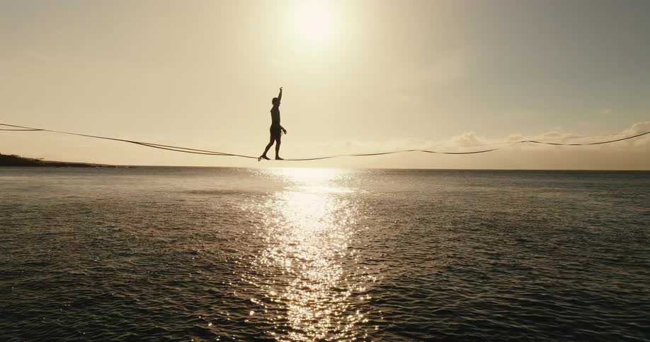 A person balances on a tightrope above the ocean during sunset