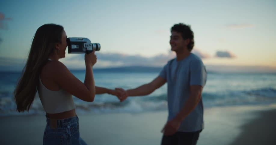 A woman holding a retro video camera is aiming it at her boyfriend on a beach