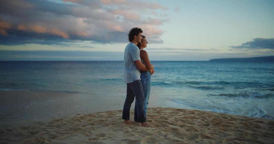 A young fully dressed couple embracing at waters edge on a beach looking at the sea
