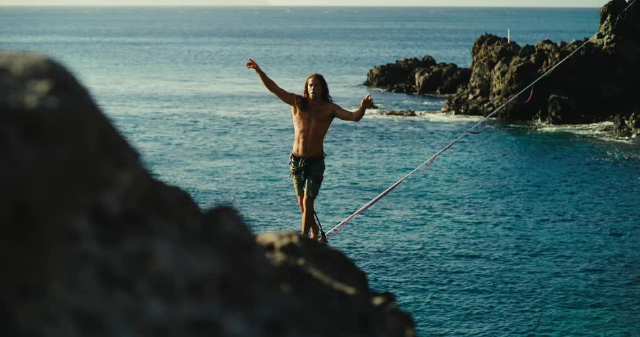 A man balances on a slackline stretched across rocks above the ocean