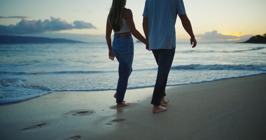 Couple holding hands walking barefoot on the beach at sunset with the sea and sunset in the background
