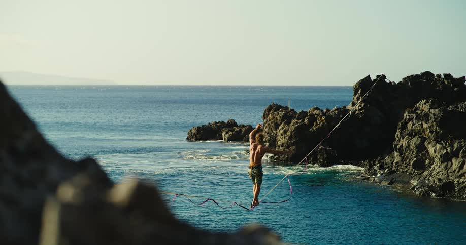 A man wearing only shorts walking on a high wire over the sea between two rock formations with a safety wire attached to his ankle