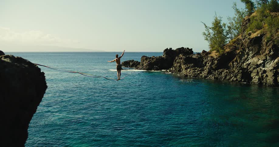A man wearing only shorts walking on a high wire over the sea between two rock formations with a safety wire attached to his ankle