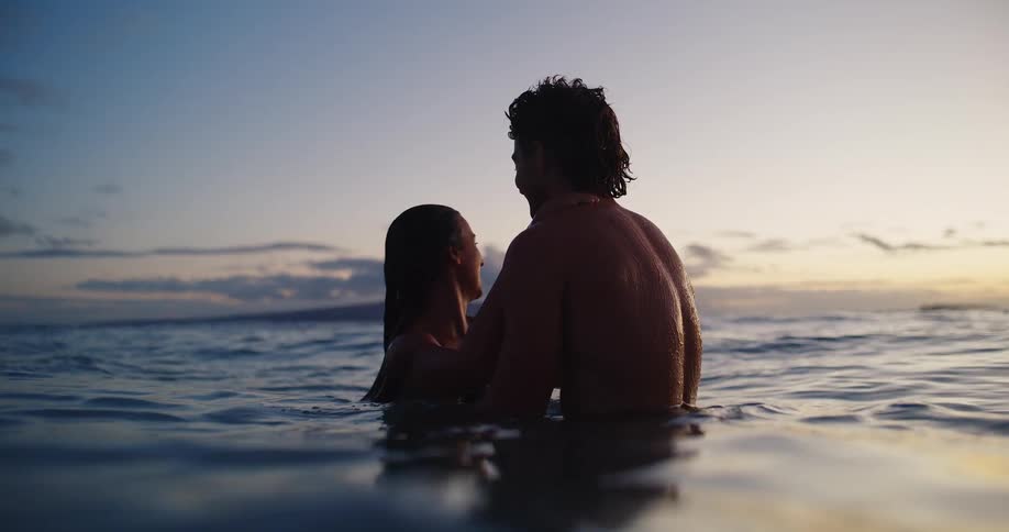 A couple is standing in the ocean at sunset with their upper bodies visible above the water they are facing each other and smiling
