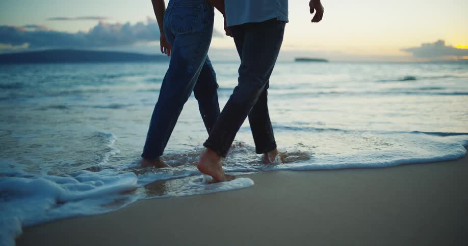 Couple walking barefoot along the seashore with their feet touched by the water at sunset