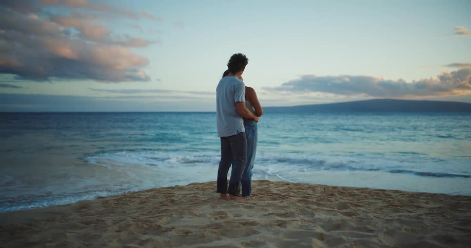 A young man holds his partner as they both stand together on the beach sand and admire the sea during daytime