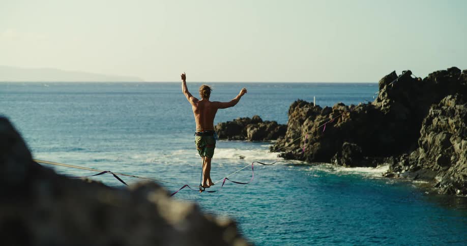 A wide shot shows a man standing on a slackline between two rocky cliffs he balances precariously above the turquoise ocean with his arms outstretched for stability