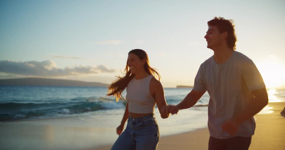 Couple holding hands runs barefoot on the beach at sunrise on a windy day