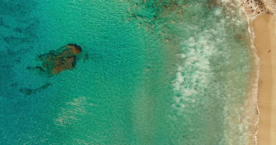An aerial view of an ocean shows a person walking on the coast of a clear turquoise body of water near a sandy beach with small waves of white foam rolling onto the shore