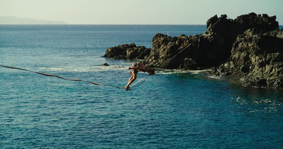 Man walks on slackline stretched above the sea between rocks and falls