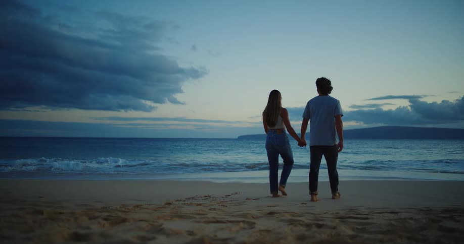 Couple holding hands walking on sand to shoreline at the beach at sunset