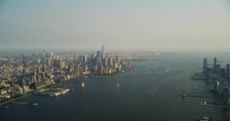 A wide static aerial shot captures the iconic lower manhattan skyline and the busy river separating it from another city on a hazy day