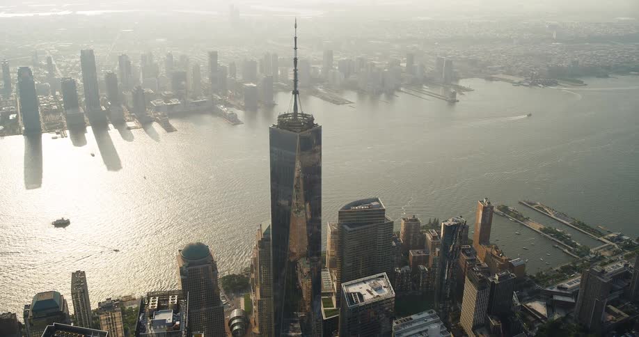 An aerial shot captures a sprawling city skyline and a large body of water possibly a harbor on a hazy day the one world trade center building stands prominently in the foreground