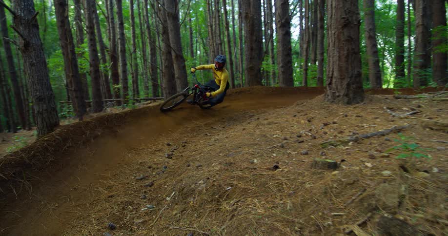 A man wearing a bright yellow longsleeved shirt and a helmet is riding a mountain bike he is shown navigating a banked turn on a dirt trail that winds through a pine forest