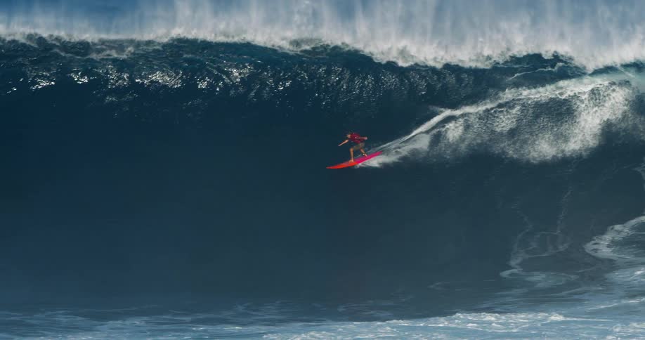 A surfer in a red shirt riding a very large powerful wave in the deep blue ocean surrounded by a dramatic crest of white spray and foam